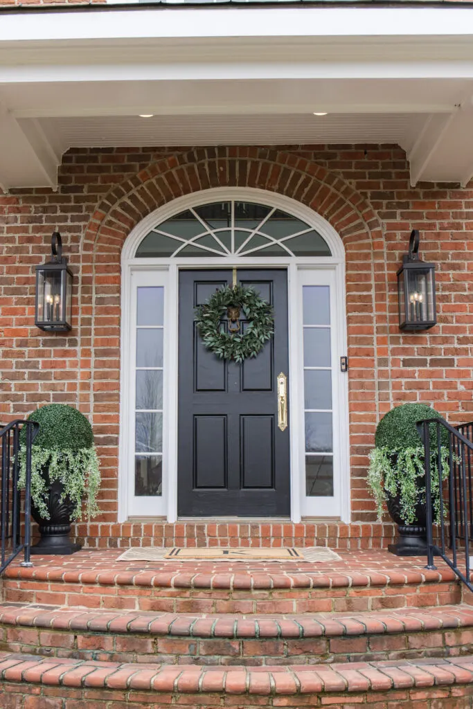 arched manor spring front porch styling, image of front porch in real life with black door, gold handles, wreath, sconces, shrubs, planters, doormat, and neutral rug