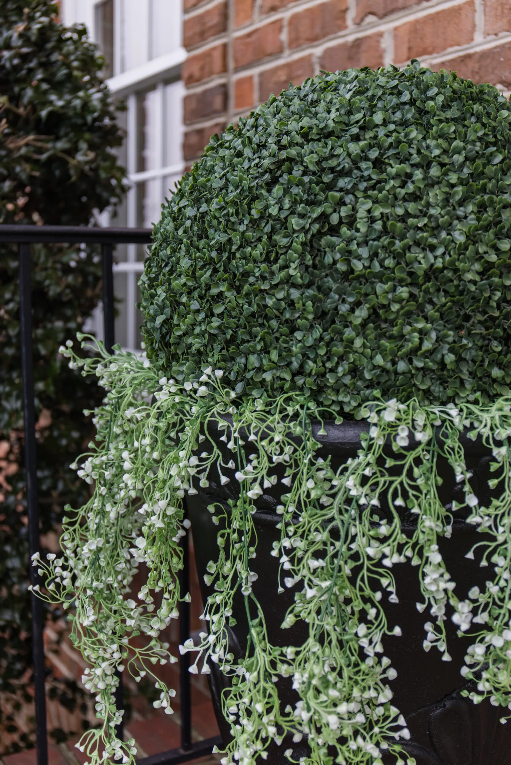Arched manor spring front porch decor, close up image of large faux shrubs ball styled with hanging plants in black decorative planter