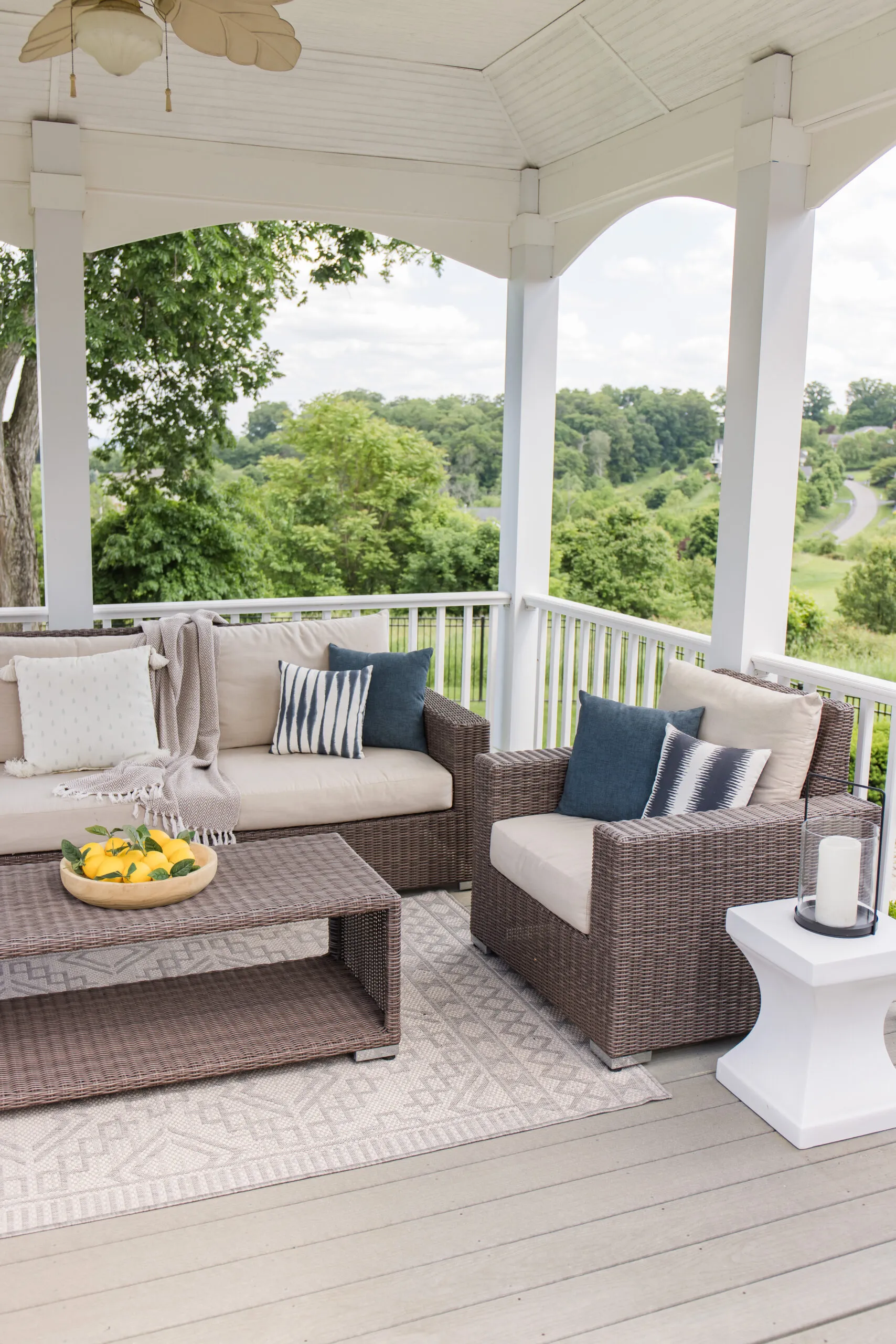 outdoor patio living space styled with wicker patio sectional and set, white accent table from amazon, neutral outdoor area rug, blue throw pillows, candle lantern, and artificial lemons in wood bowl