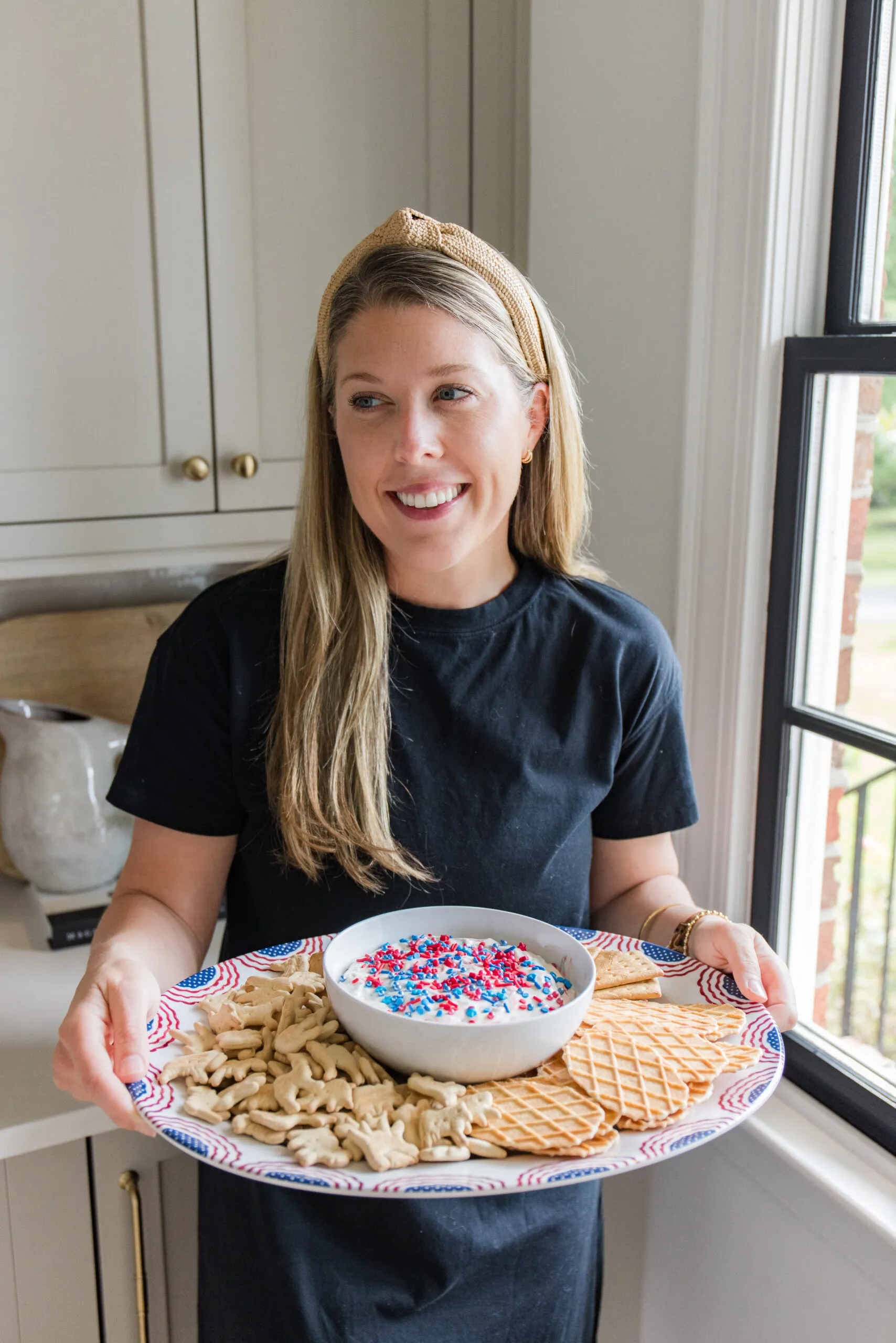 megan wearing a navy blue dress holding a serving platter with snacks and a white bowl filled with dip